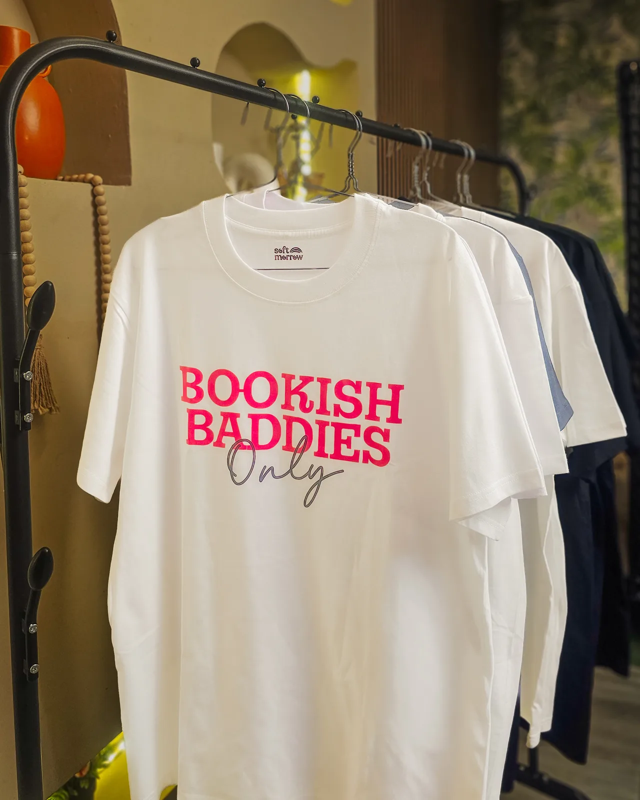 Softmorrow tees on a rack, featuring the Bookish Baddies Only tee, against a warm niche-shelf wall